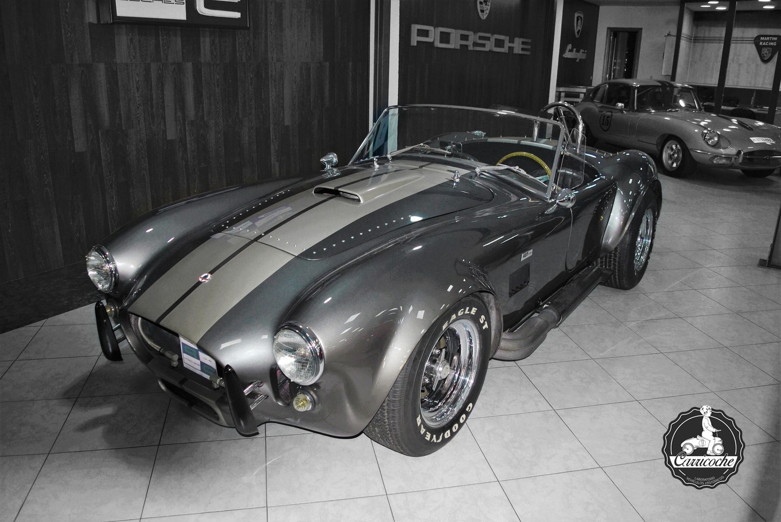 Recreación de un Shelby Cobra de color gris con franjas, expuesta en el interior de una colección junto a un Jaguar E-Type y cartelería de marcas deportivas.