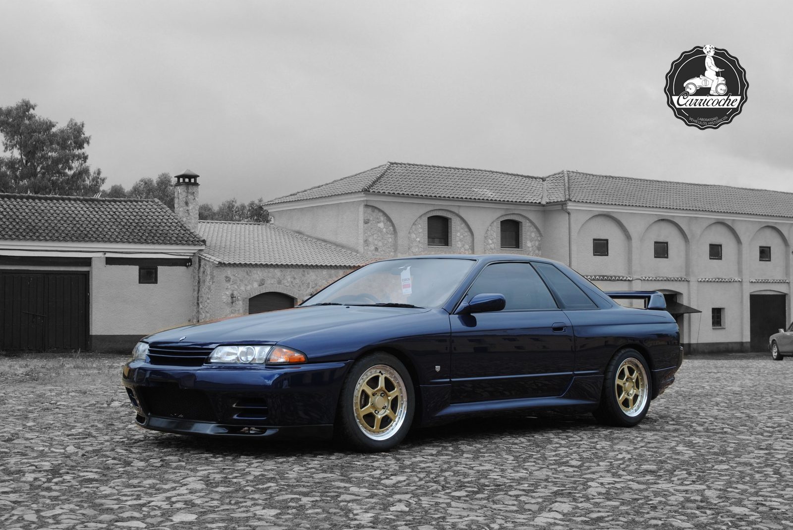 Nissan Skyline GT-R R32 de 1991 de color azul oscuro, con llantas doradas, sobre un empedrado frente a edificaciones rurales.